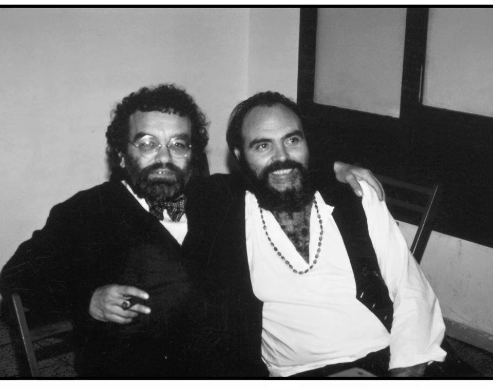 Dos hombres con barba sentados juntos y sonrientes, en blanco y negro: Fernando Arrabal, con gafas redondas y un puro, y Ángel Alonso